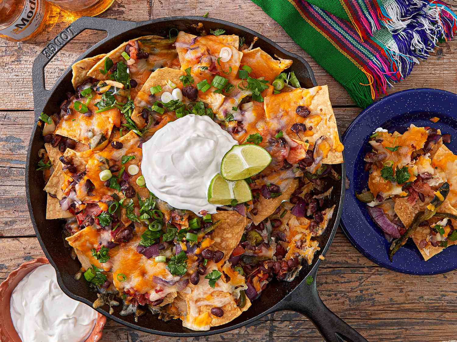 A skillet dish of loaded nachos garnished with lime wedges and sour cream; a small plate with a serving, on a wooden table
