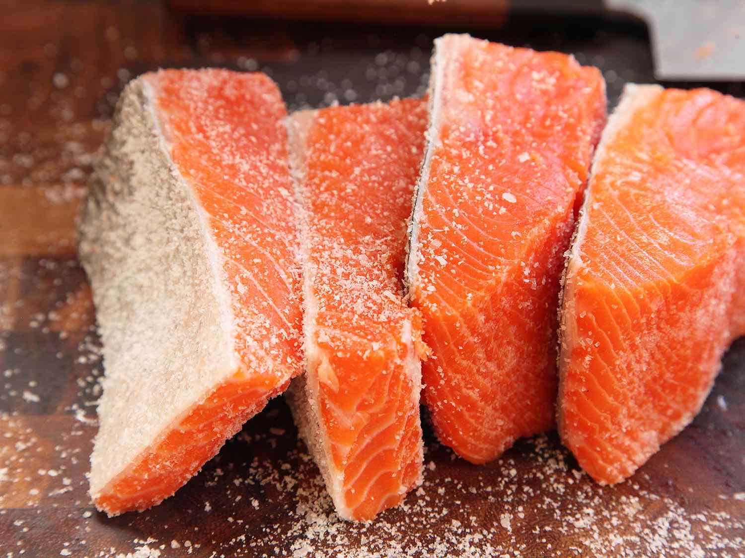 Four salmon fillets on a wooden cutting board, seasoned with kosher salt, before being cooked sous vide.