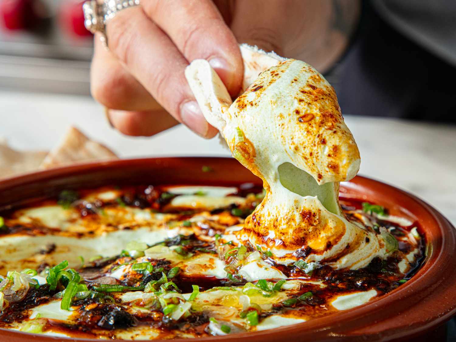 A hand dipping a piece of bread into a baked cheese and herb dish served in a terracotta bowl