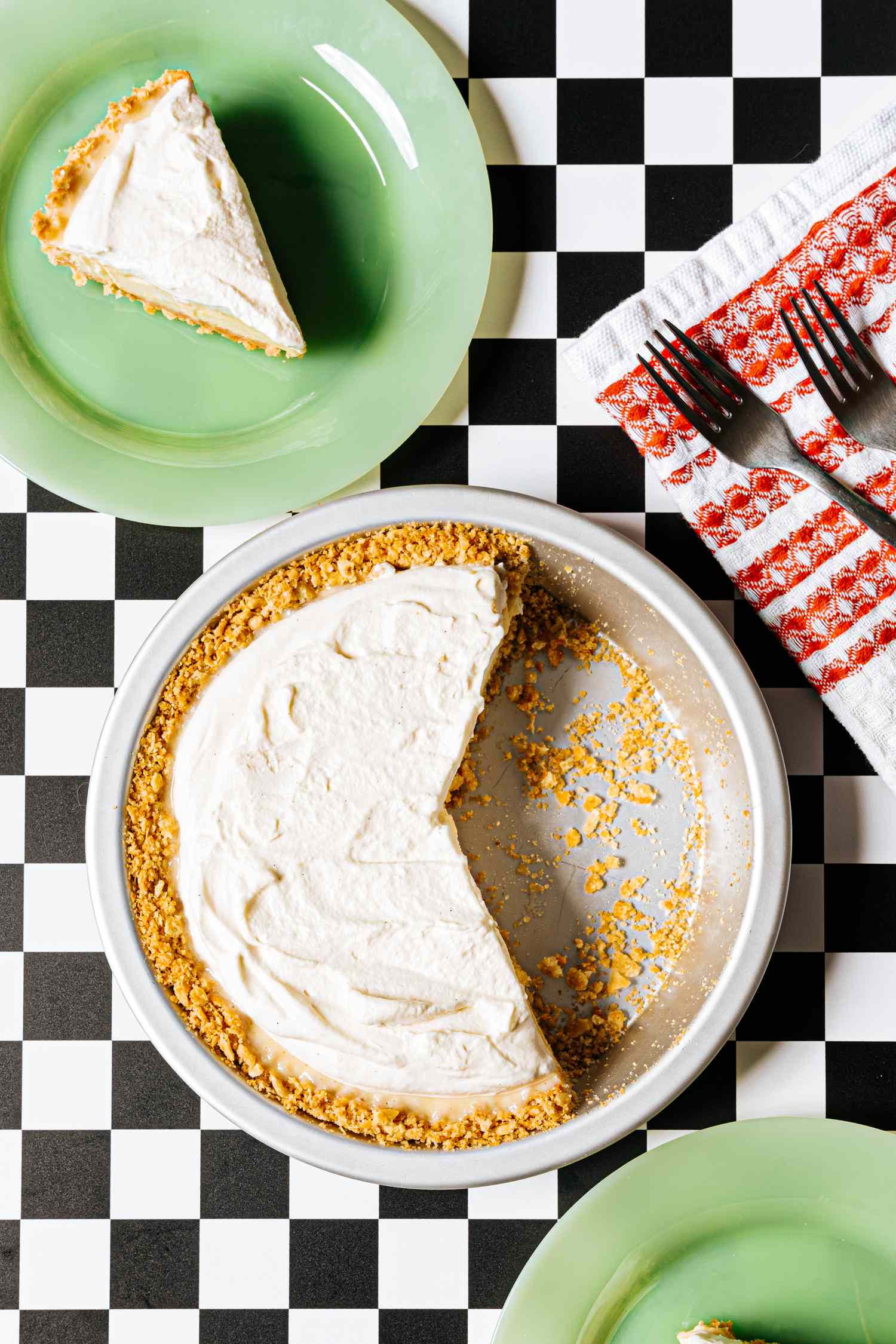 A partially eaten pie on a checkered surface with a slice served on a green plate nearby accompanied by a red napkin with two forks
