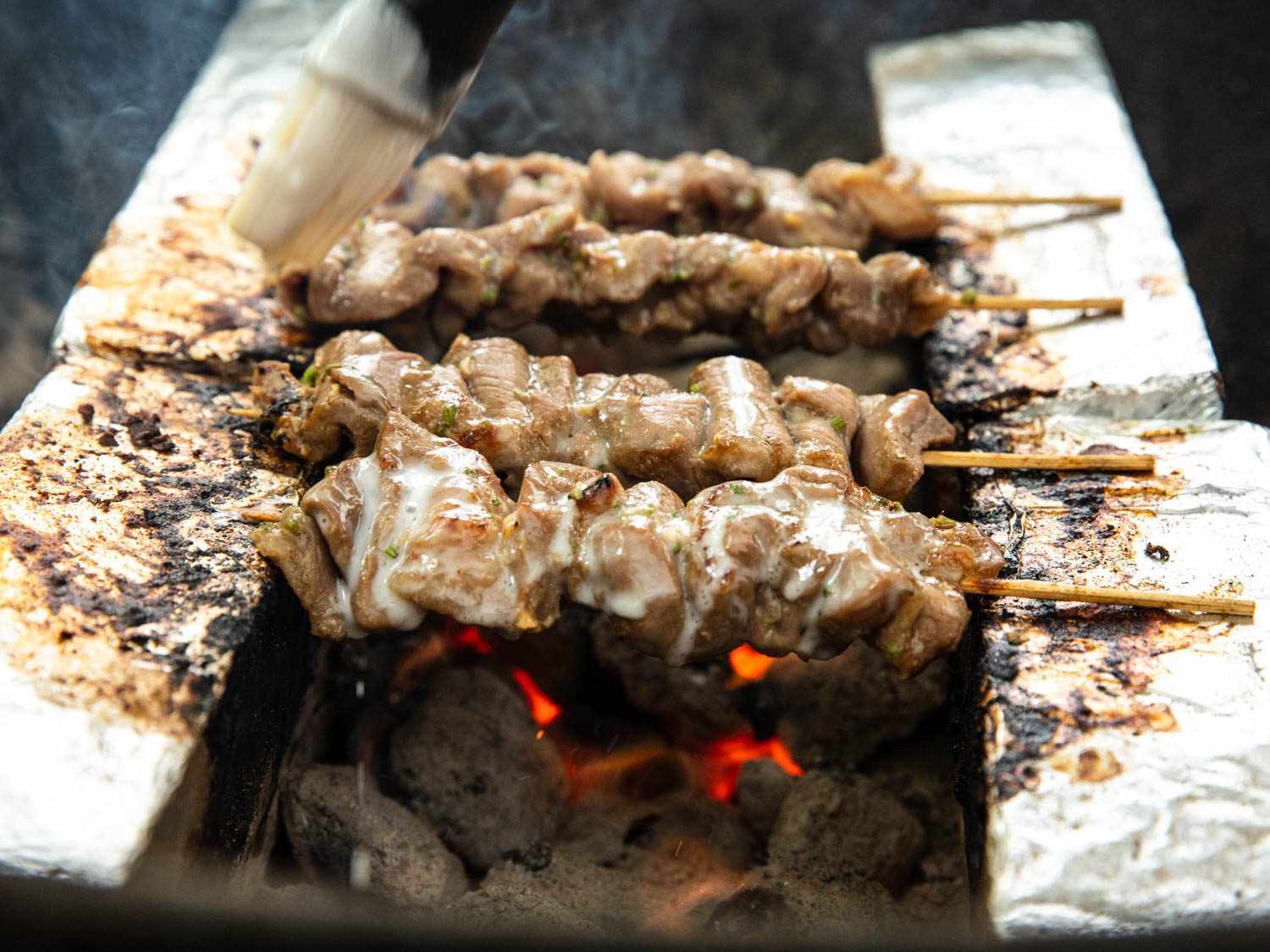 Several Thai pork skewers cooking over charcoal, suspended between two parallel rows of foil-wrapped bricks