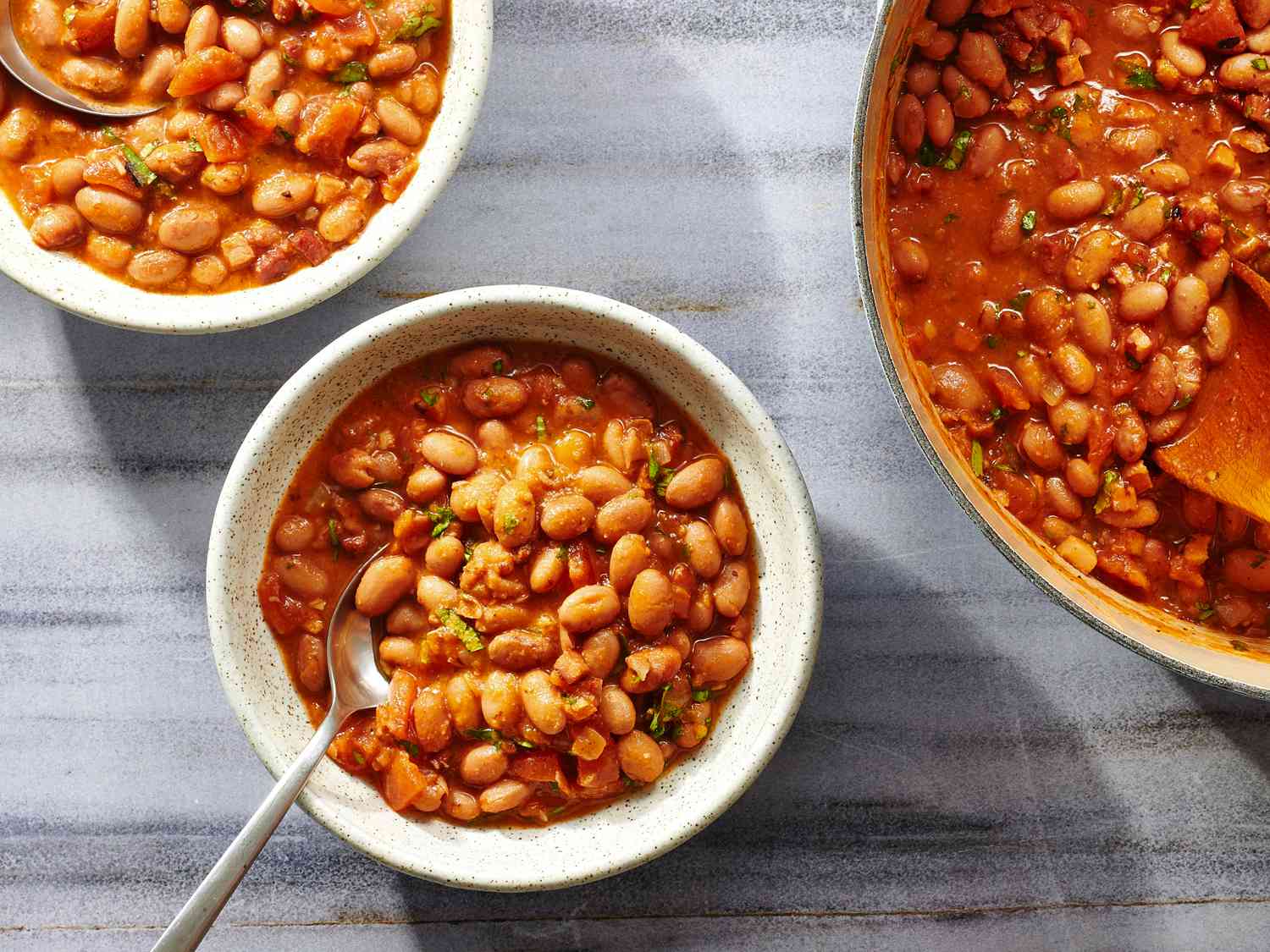 Two bowls and a pot of frijoles charros, on a stone background.