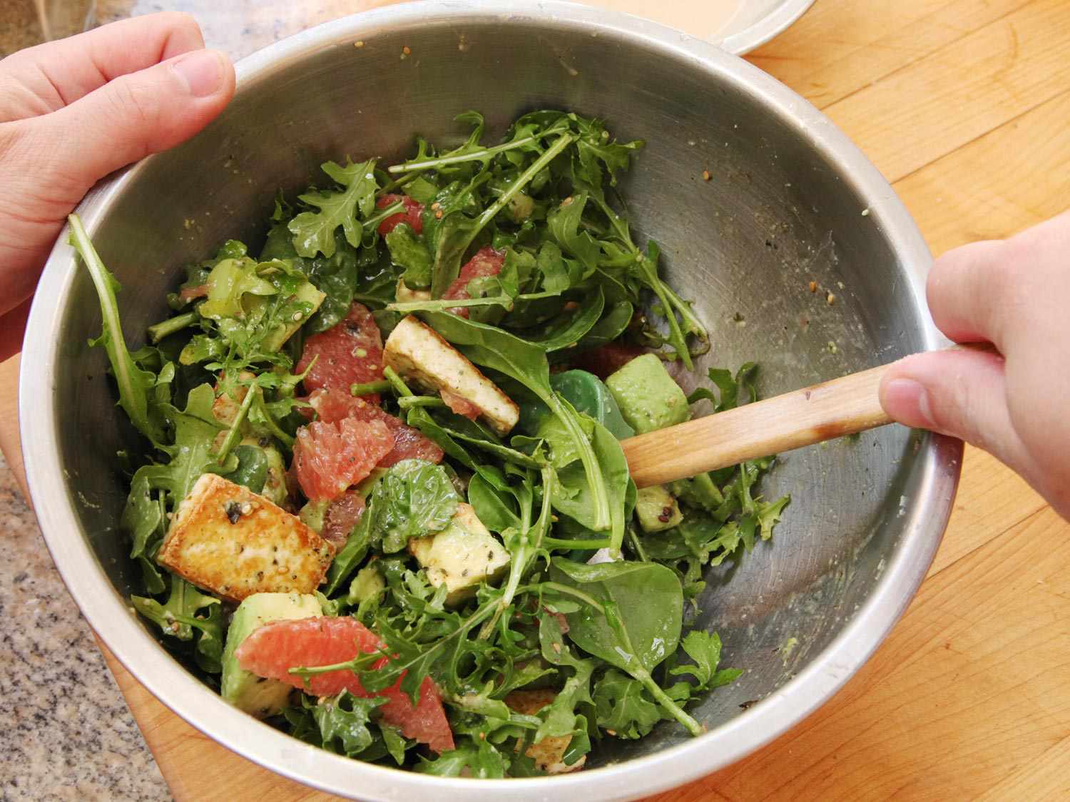 Tofu, avocado, and grapefruit in a metal bowl being tossed with greens.