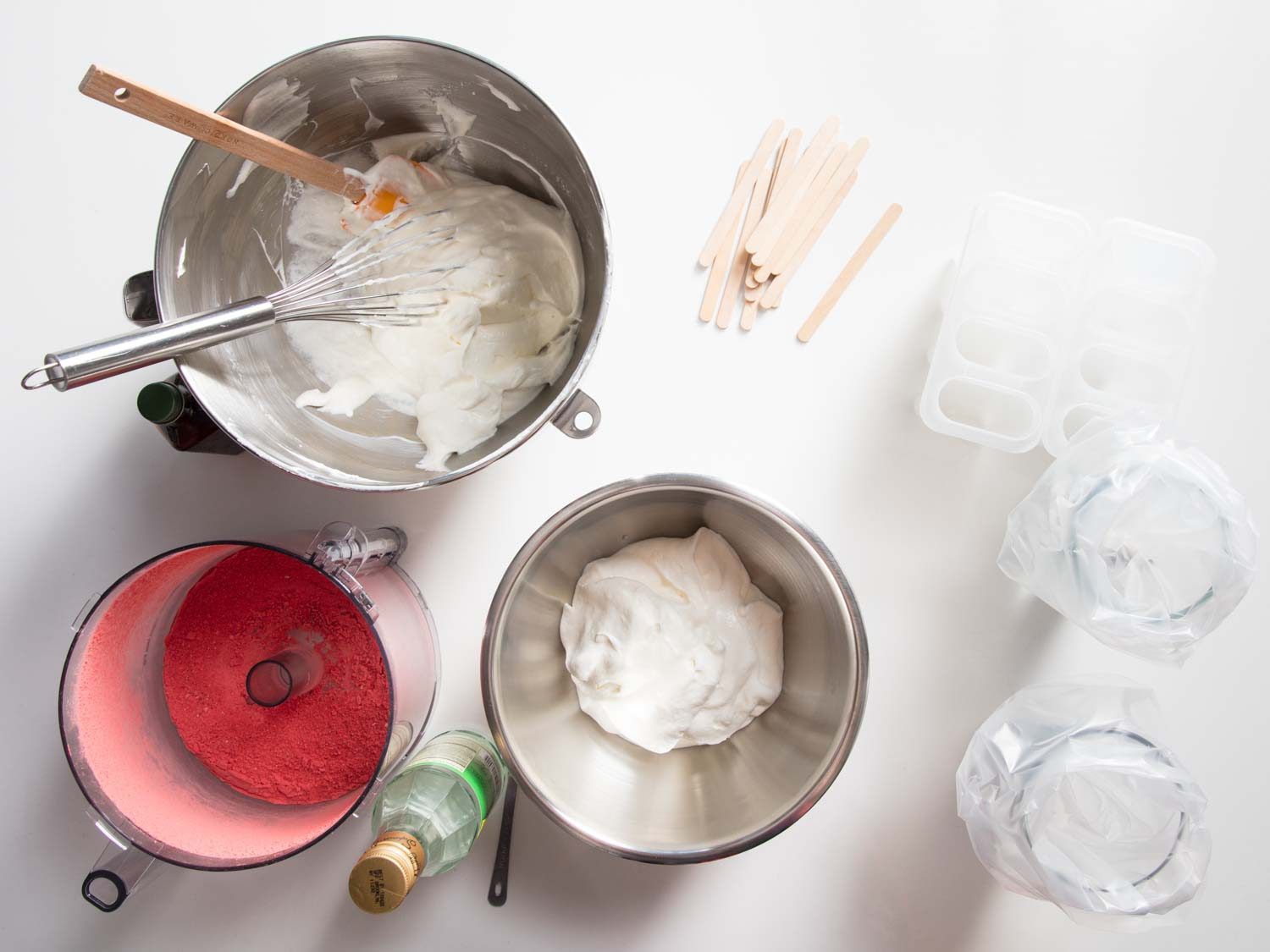Top view of a large mixing bowl of Swiss meringue next to a processor containing powdered freeze dried strawberries and a smaller mixing bowl of Swiss meringue. On the table are also a bottle of rose water, popsicle molds, and two glasses holding piping bags.
