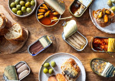 overhead shot of an array of tinned fish, or conservas, on a wooden table with bread and olives.