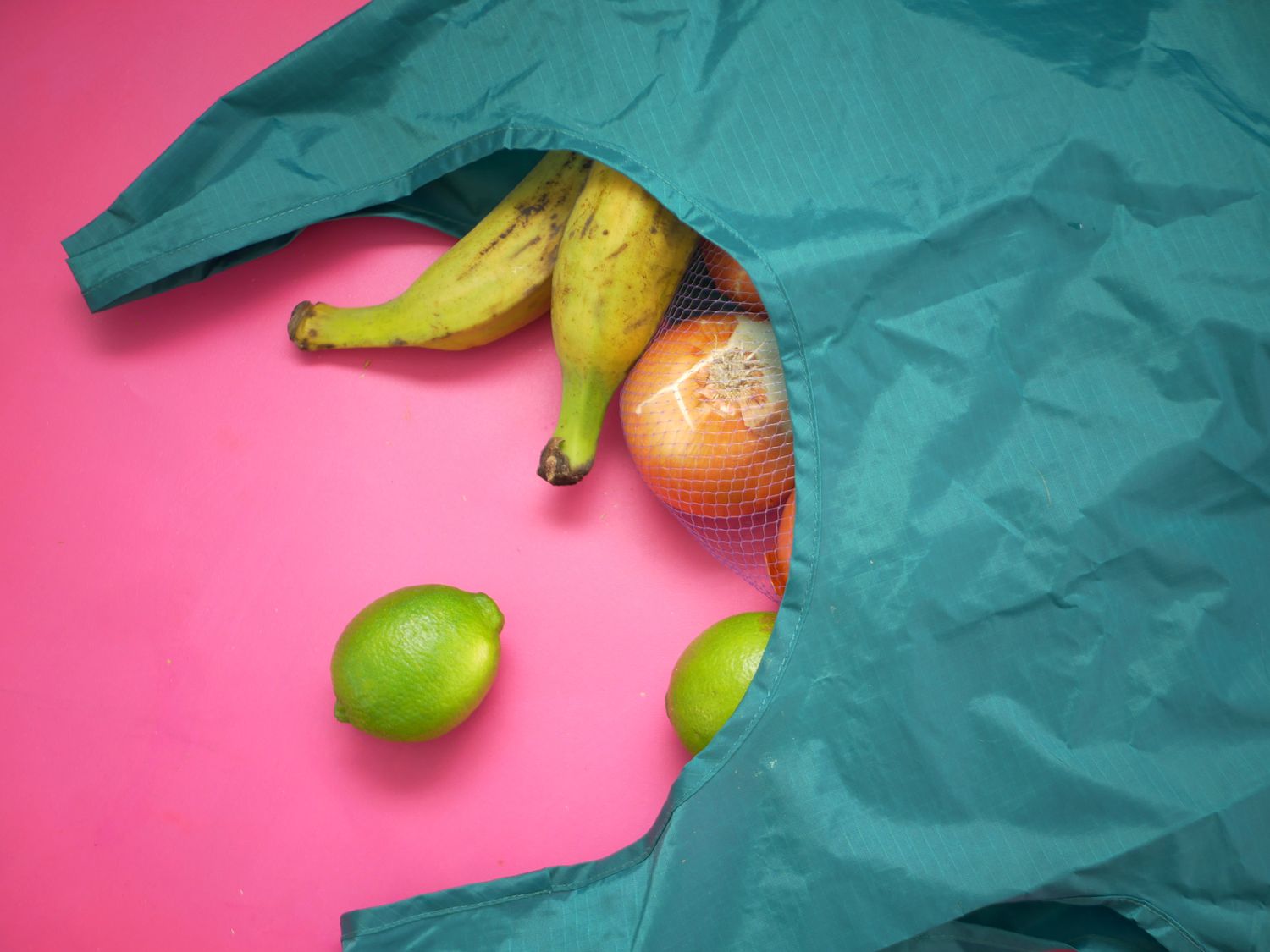 A dark teal Baggu grocery bag on its side with plantains, onions, and limes spilling out
