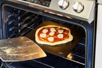 A person using a pizza peel to place a pizza onto a baking steel.