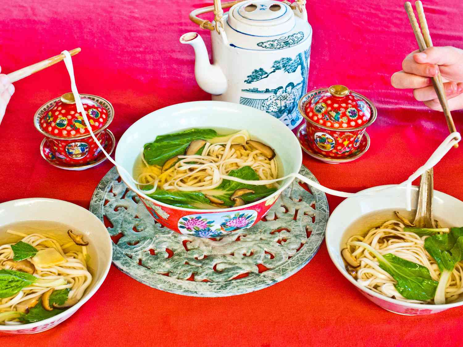 A large bowl of longevity noodles are served on a red table, alongside a pot of tea. Diners serve themselves from the bowl, transferring the long noodles with chopsticks.