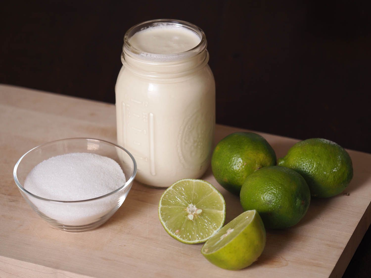 A glass bowl of sugar, pint mason jar of cream, and fresh limes, one cut in half.