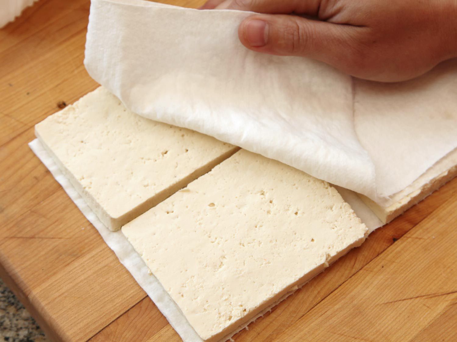 Thin slabs of tofu are dried between paper towels on a cutting board.