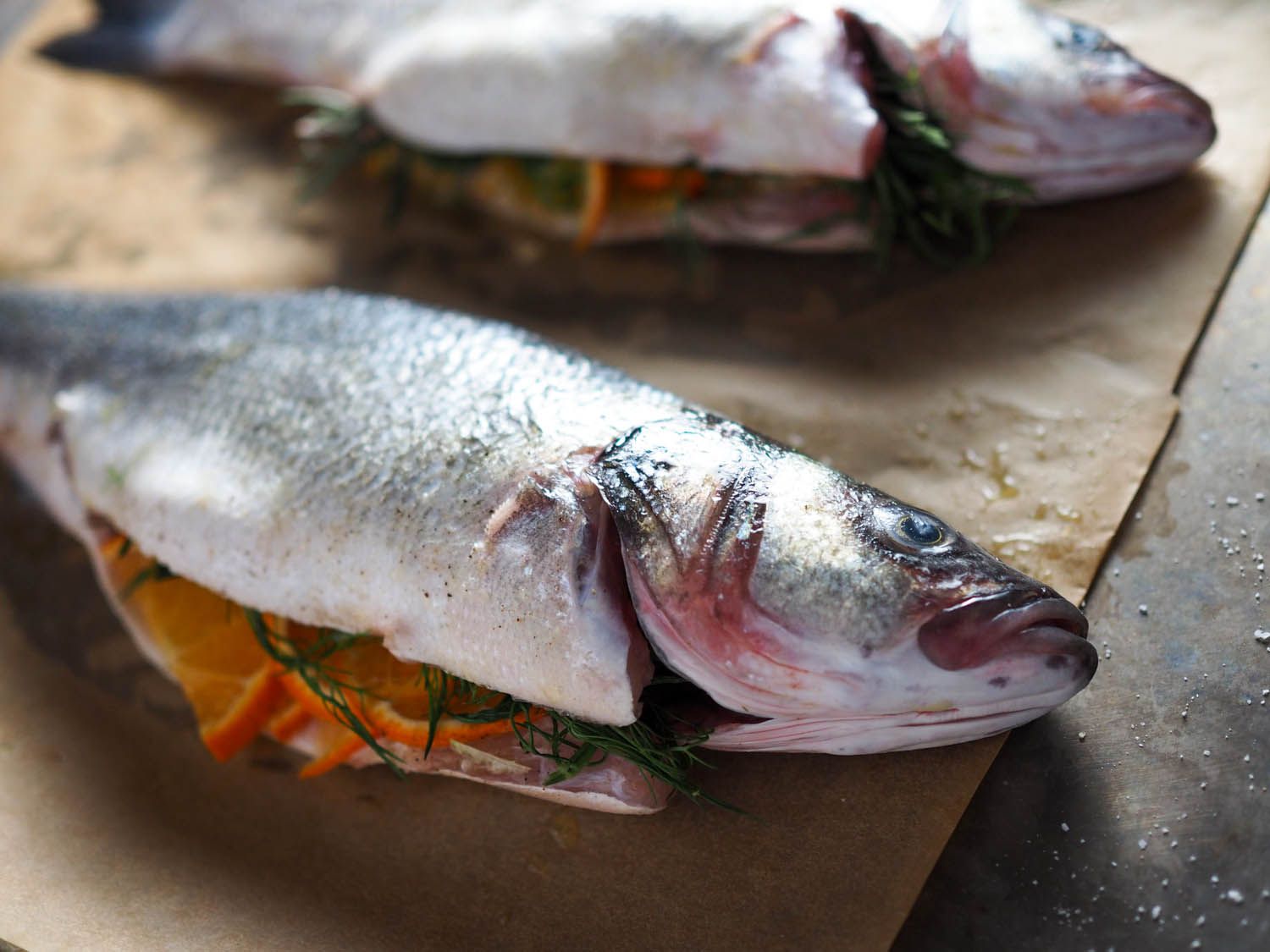 A close-up of two whole branzini, their bellies stuffed with fennel fronds, rosemary, and tangerine slices, on brown parchment paper. 