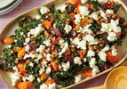 A salad made with kale roasted root vegetables and crumbled cheese topped with nuts on a rectangular serving dish