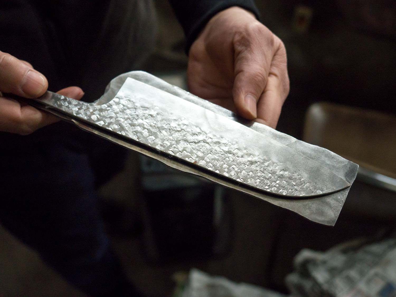 A finished santoku blade is held against a piece of raw steel.