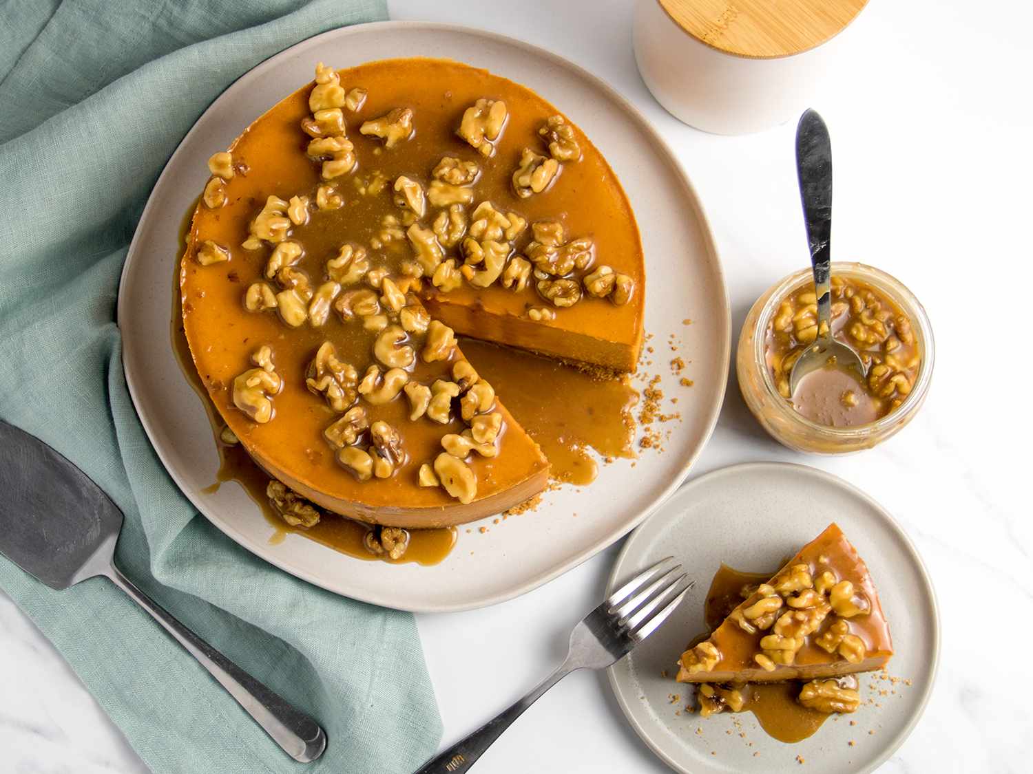 A deep orange sweet potato cheesecake drizzled with a flood of walnuts and caramel sits on a large plate with one slice missing; that slice is on a serving plate next to it.