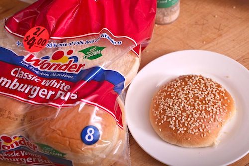 Image of a pack of Wonder Bread brand hamburger buns. Nearby is a plate with a sesame-encrusted top bun.