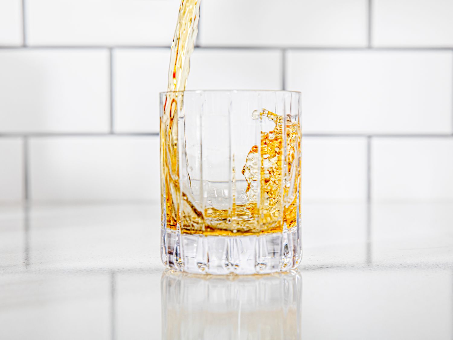Whiskey being poured in a rocks glass in front of a white kitchen backsplash