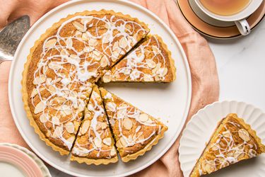 A bakewell tart served on a plate, sliced with one piece on a separate dish next to a cup of tea