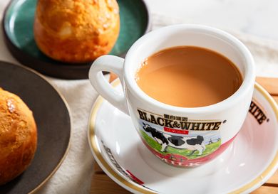 Cup of milk tea in a logoed mug on a plate with baked goods in the background