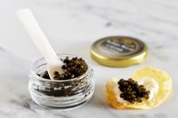 An open jar of Olma kaluga royal caviar next to a potato chip topped with crème fraîche and a scoop of caviar