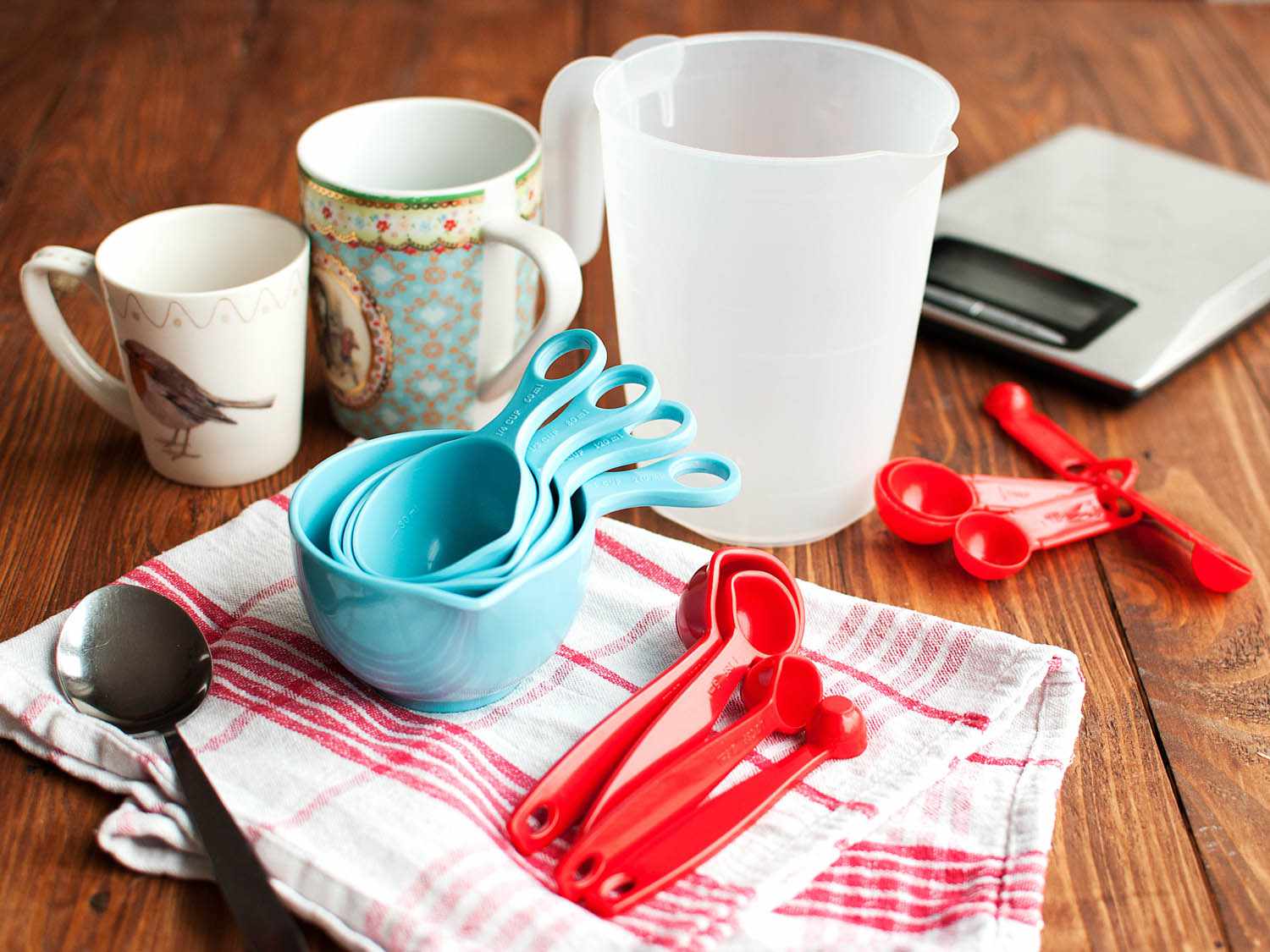 Measuring spoons, measuring cups and a kitchen scale, along with a tea towel on a wooden table. 