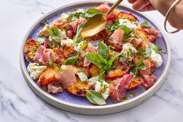 A hand drizzling dressing over a grilled peach salad with prosciutto and basil
