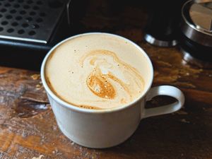 A cup of coffee on a wooden table with foam art on the surface, a coffee machine partially visible in the background