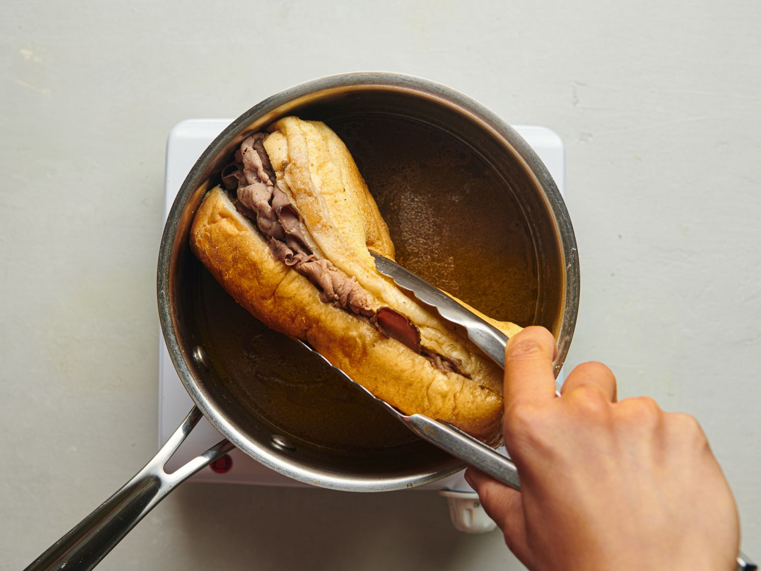 A pair of tongs dipping a meat-filled sandwich into the saucepan of liquid. This is the preparation method for if you want a "dipped" version of the sandwich. After the sandwich is dipped it would be topped with roasted peppers and giardiniera. 