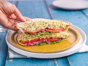 A hand holding a chickpea salad sandwich cut in half, filled with lettuce, red onion, and tomato, placed on a plate