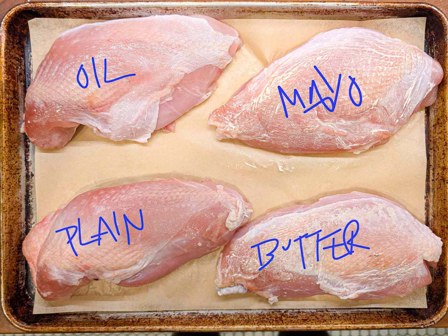 Four raw turkey breasts labeled oil mayo plain and butter on a baking sheet lined with parchment paper