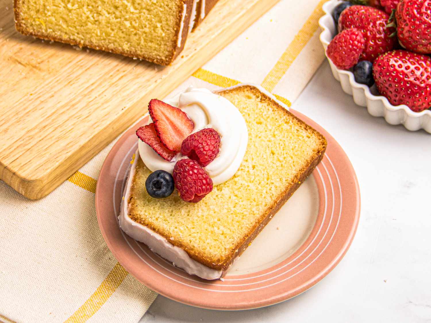 Slice of pound cake topped with whipped cream and mixed berries on a plate, wooden board and bowl of berries in the background.