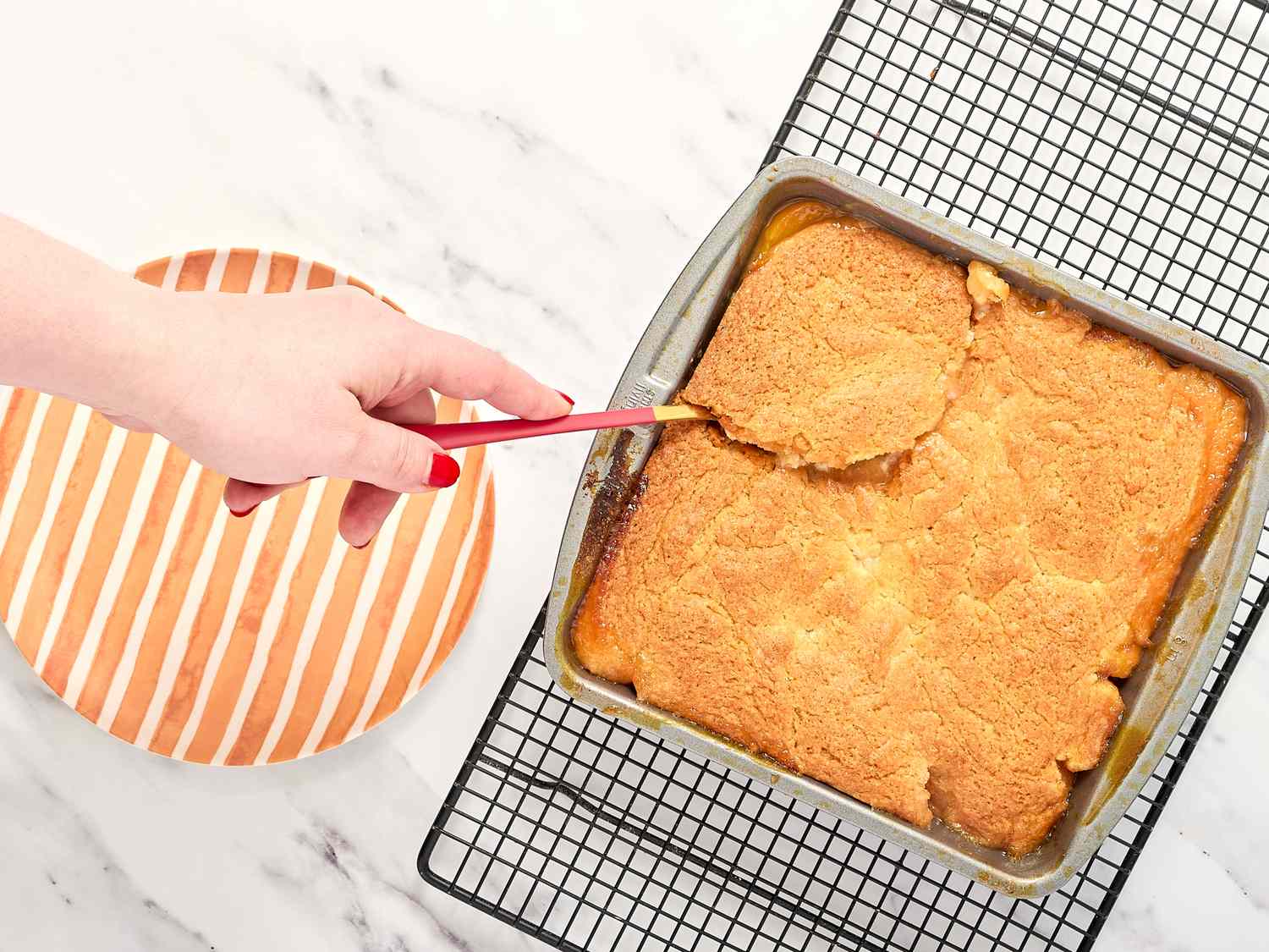 A hand holding a utensil scooping from a baking dish containing a peach cobbler on a cooling rack