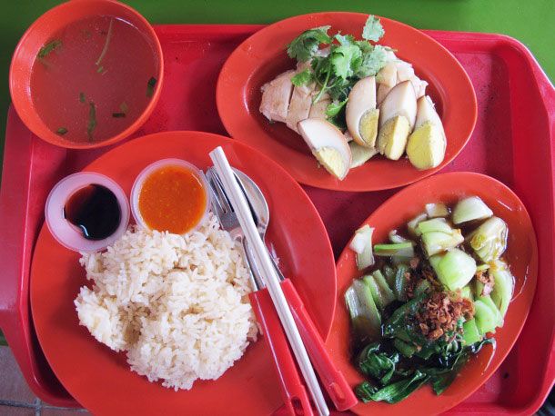 A Hainanese chicken and rice plate with broth, sauces, and a side of simmered bok choy topped with browned shallots.