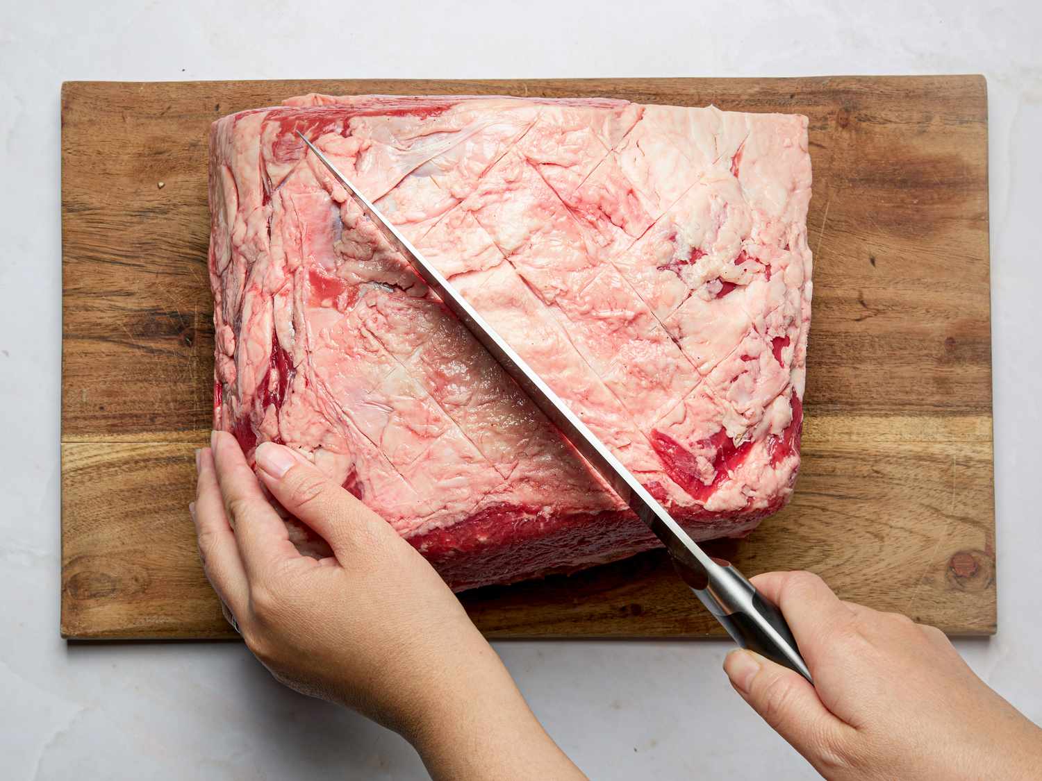 Person scoring the fat on a raw prime rib on a wooden cutting board with a sharp knife