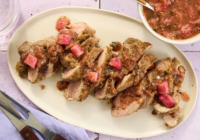 sliced pork with rhubarb sauce drizzled on it, on aa cream colored plate, with bowl of rhubarb sauce to the side, on a tile surface.