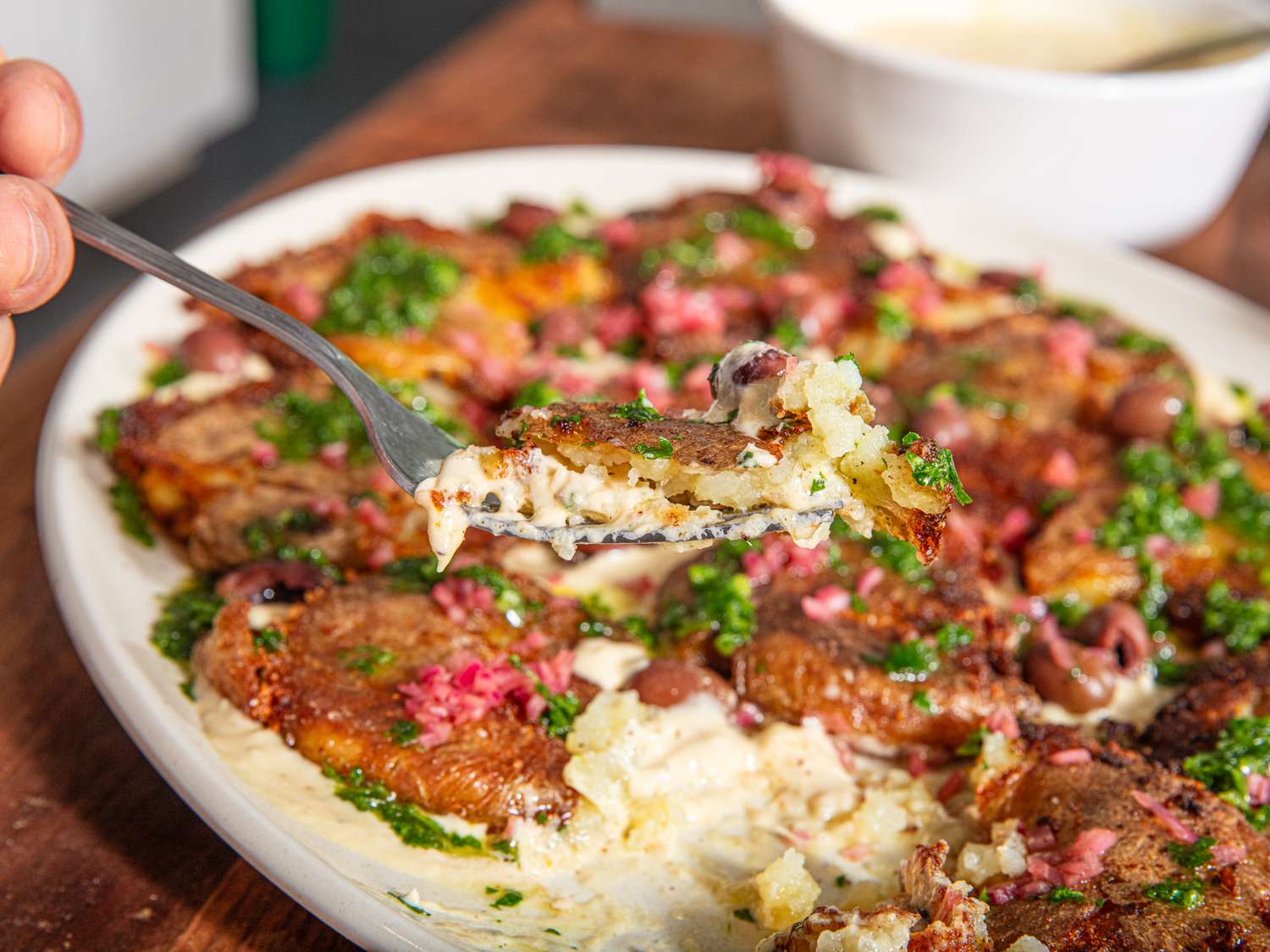 A serving of smashed double sauce potatoes on a fork, with a plate full of the dish in the background