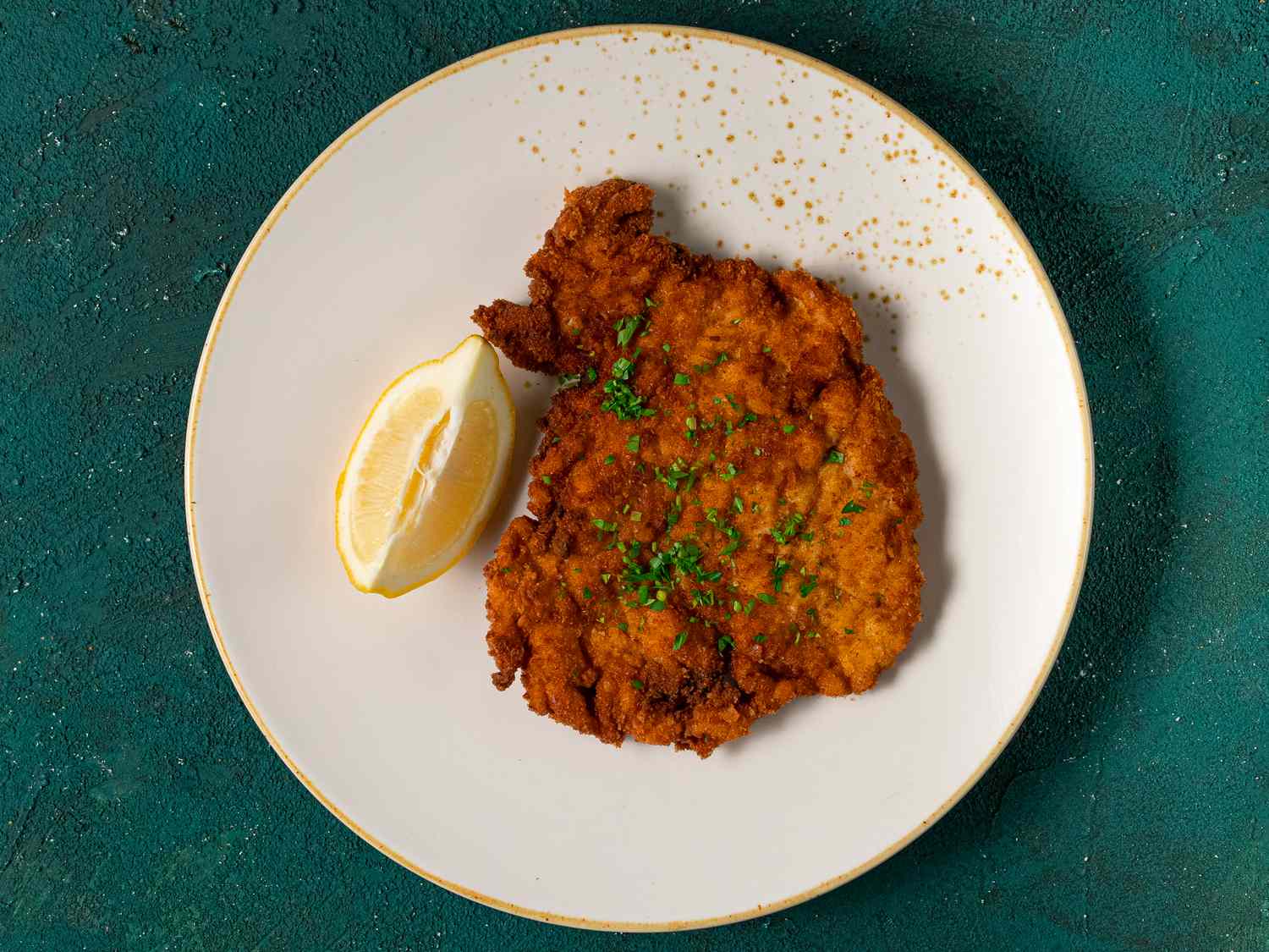 A golden brown fried chicken schnitzel cutlet with a lemon wedge on a gold and white ceramic plate on a teal-colored textured background.