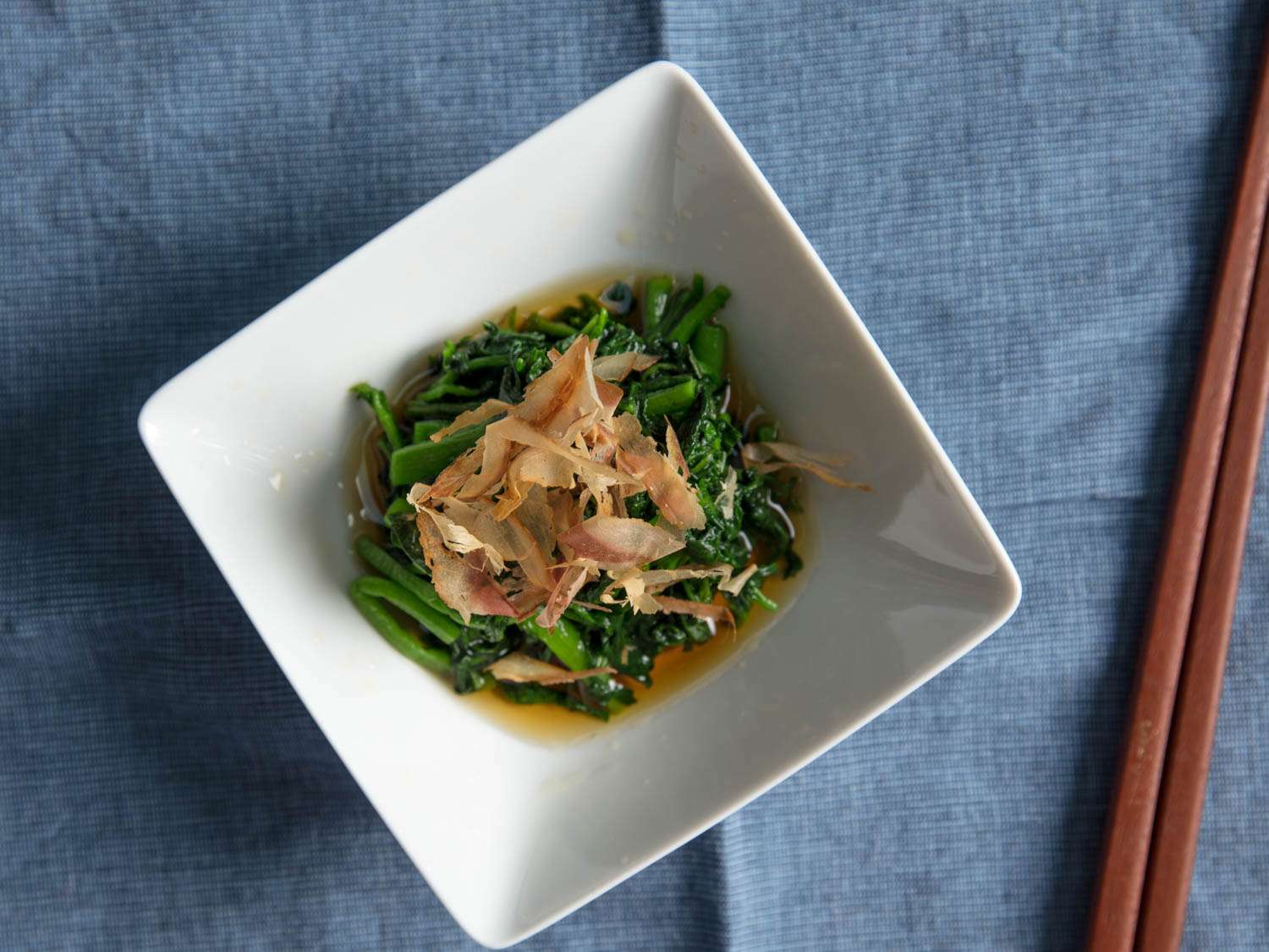 Overhead shot of a dish of ohitashi topped with shaved bonito and a pair of chopsticks on the side.