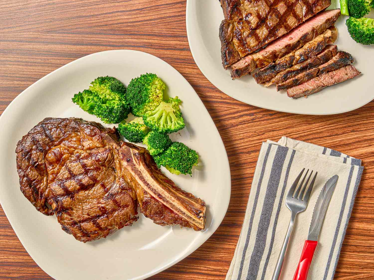 Two plates with steak and broccoli one steak cut into slices and a striped napkin with cutlery on the side
