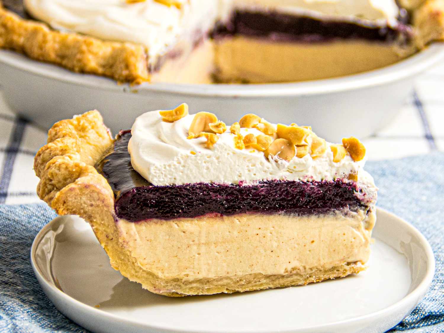 A slice of peanut butter and jelly pie topped with whipped cream and peanuts on a plate pie dish in the background