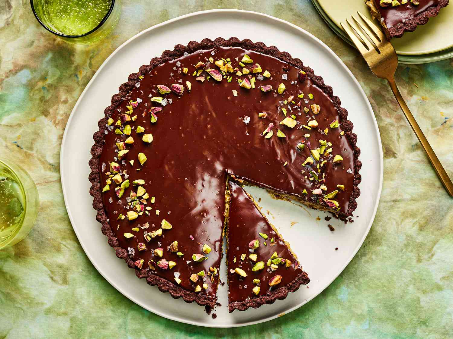 Dubai Chocolate Tart topped with pistachio, on green tablecloth with 2 glasses of water, and stacks of plates in the corner
