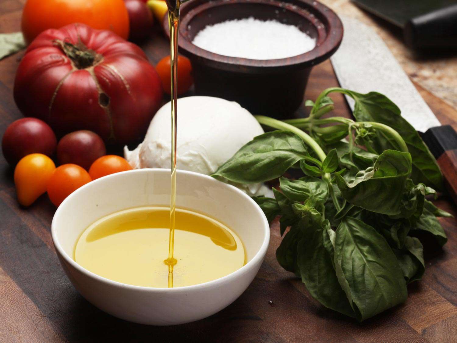 Olive oil being poured into a bowl with Caprese salad ingredients around it.