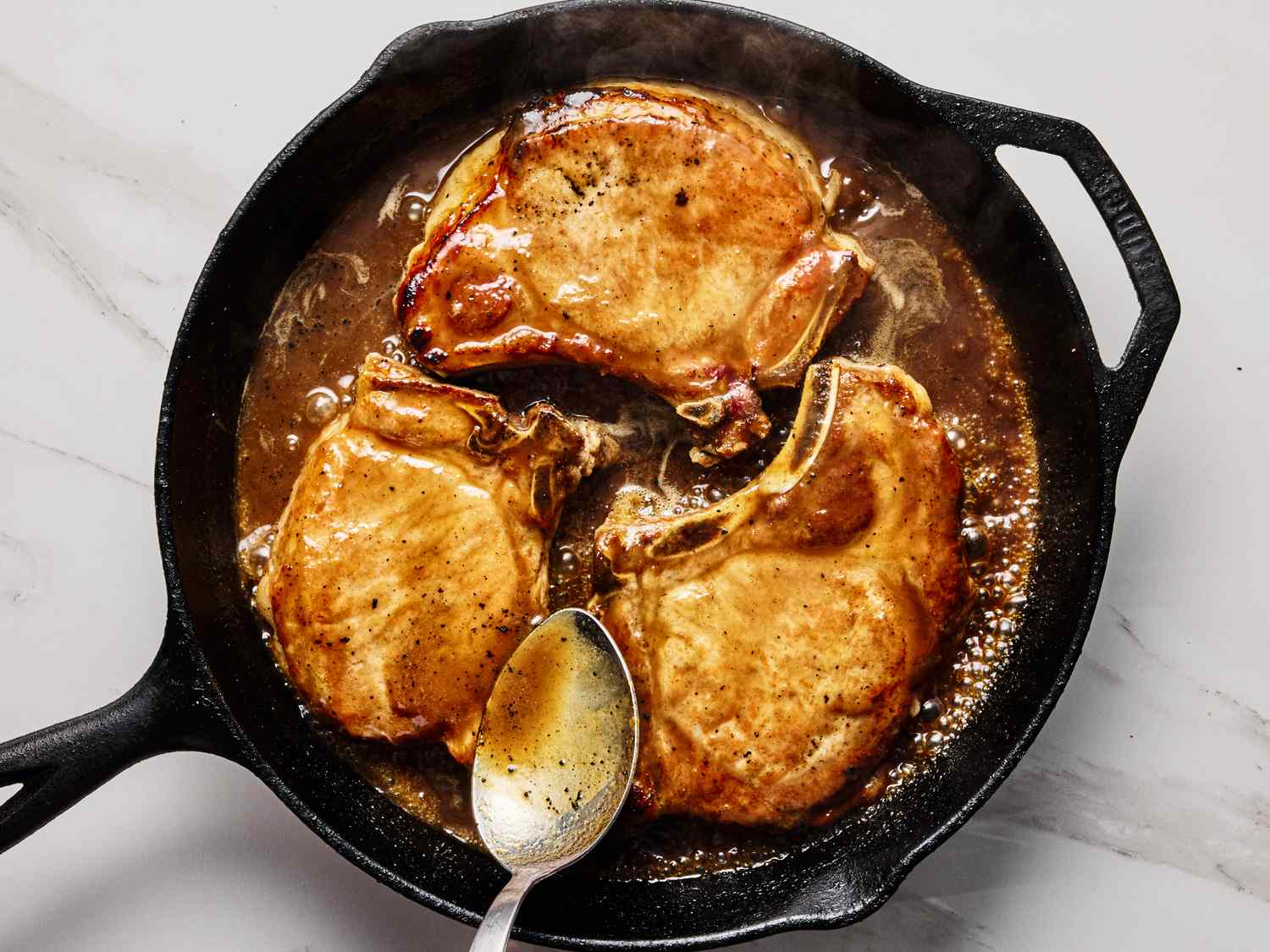 Pork chops cooking in a skillet with sauce spoon resting in the pan