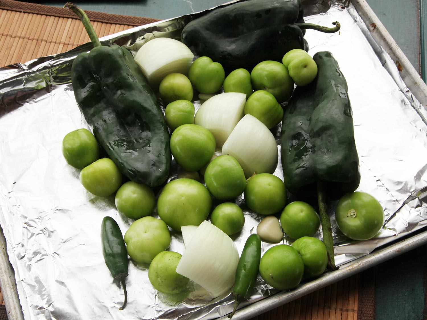 A foil-lined baking sheet with whole poblanos, serranos, and tomatillos along with some peeled garlic and quartered onion.