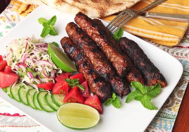 A platter of Pakistani Seekh kebabs with slaw, cucumbers tomatoes, and lime wedges.