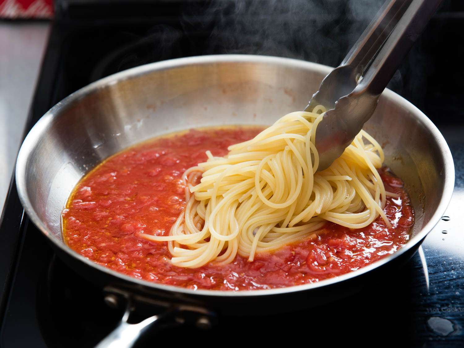 Using tongs to place cooked spaghetti into a red pasta sauce cooking in a skillet.