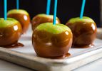 Caramel-dipped Granny Smith apples with blue sticks on a baking tray.