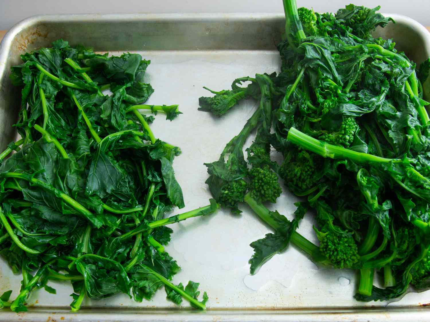 Side-by-side piles of blanched broccoli rabe leaves and stems on a rimmed baking sheet.