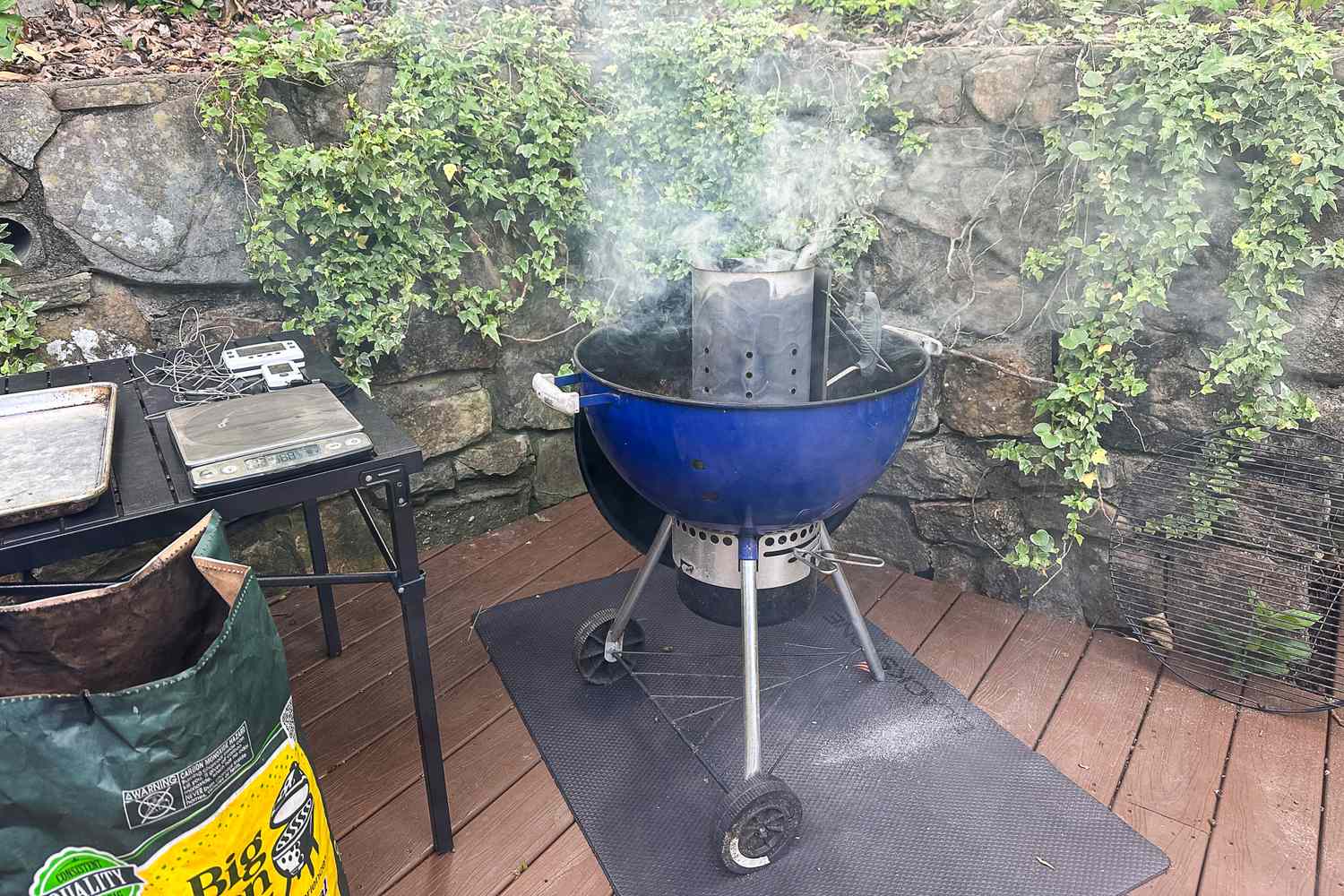 A charcoal grill with a chimney starter emitting smoke situated on a wooden deck near ivycovered stone walls with grilling supplies nearby
