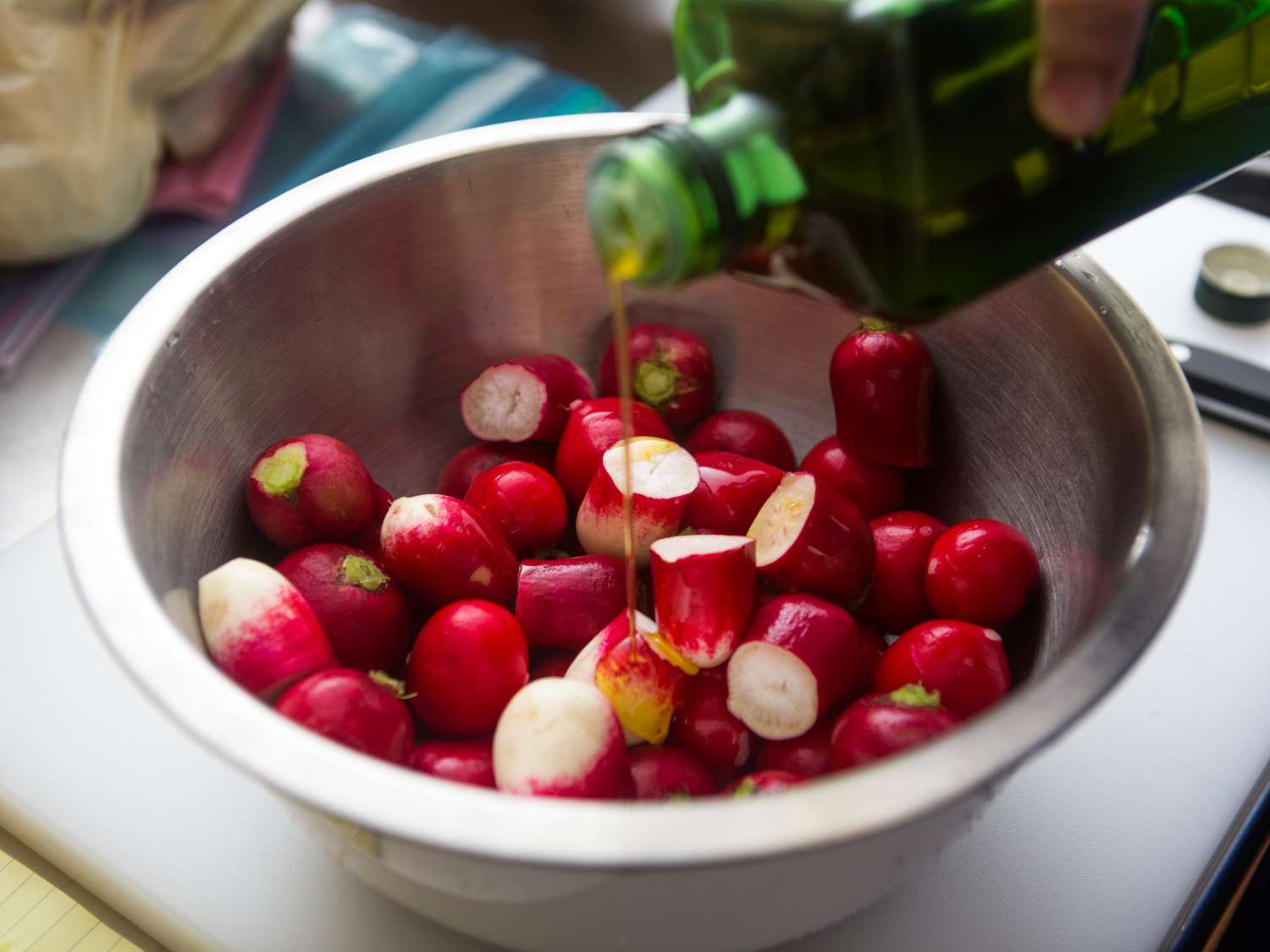A mixing bowl containing topped and tailed radishes. The larger ones have been halved. The author is drizzling them with olive oil.