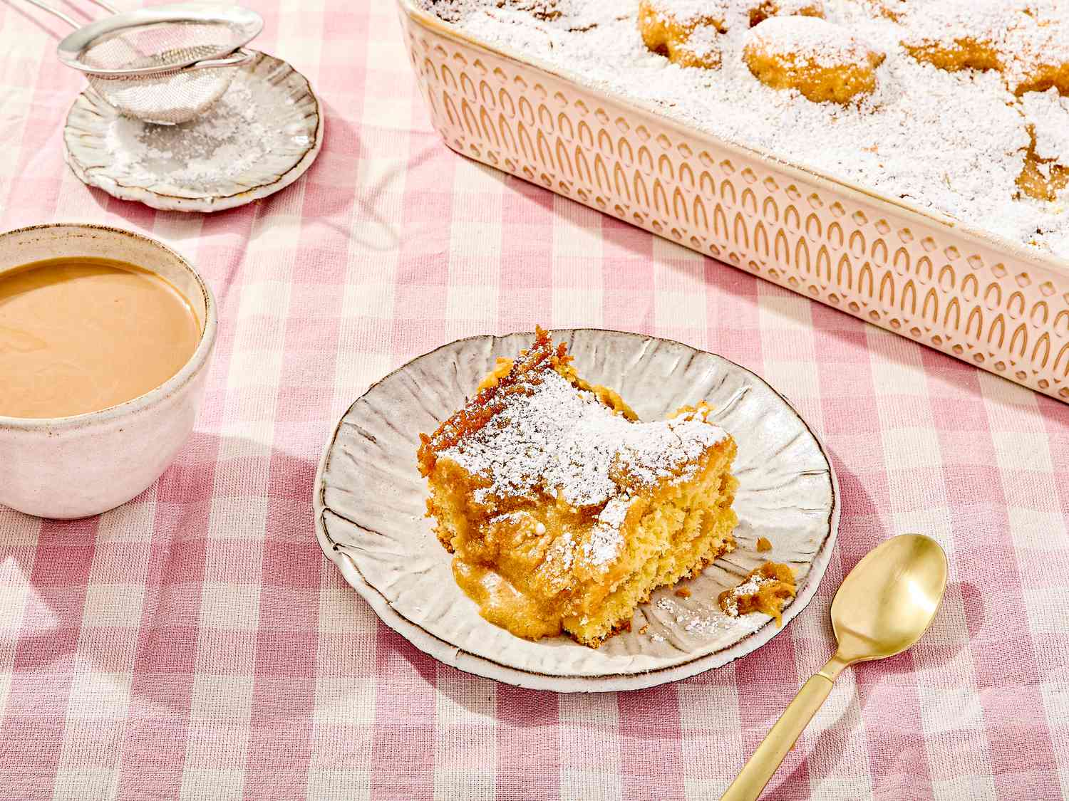 A piece of gooey butter cake on a plate with powdered sugar, with a cup of coffee and a serving tray of cake in the background
